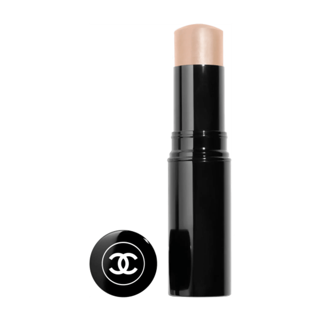 A black Chanel makeup stick with a beige-colored product exposed. The stick stands upright with its round black cap, featuring the white Chanel logo, placed nearby on its side.