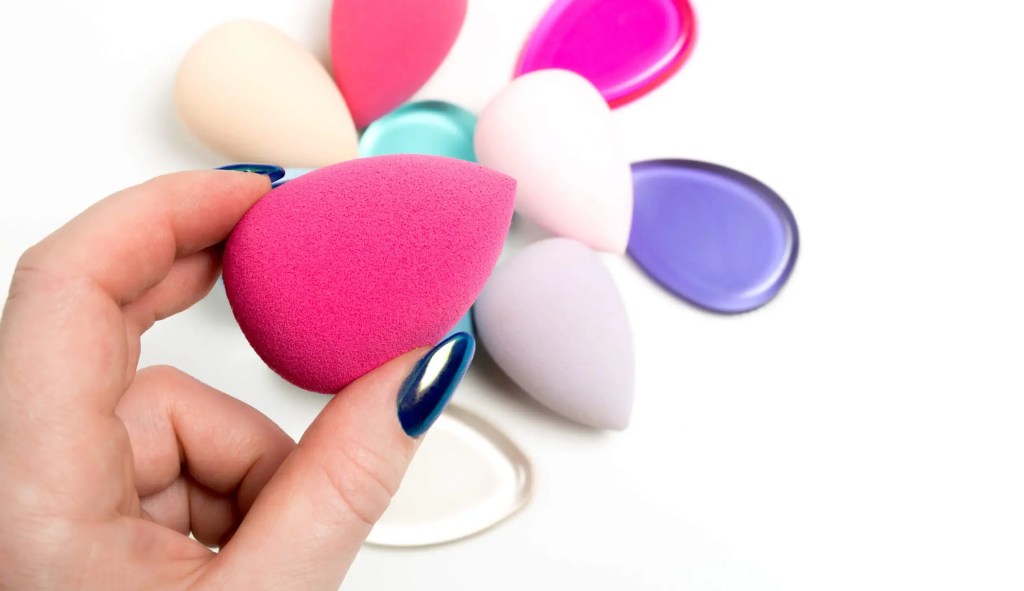 Hand holding a pink makeup sponge with colorful beauty blenders in background — perfect for applying foundation evenly on mature skin.