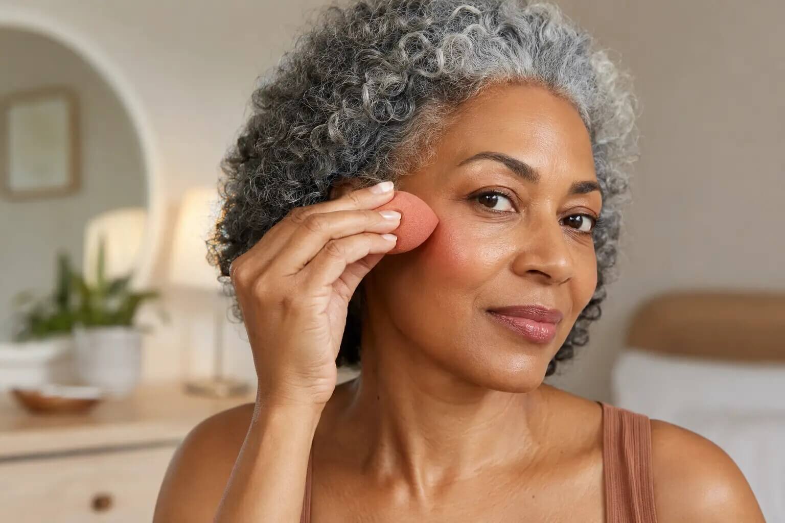 A mature woman blending cream blush upward with a damp sponge